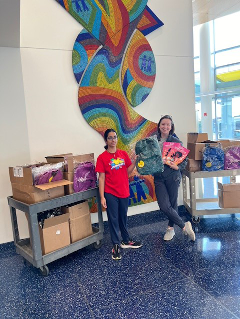 A young girl with dark hair and glasses in a red shirt holding up a backpack. Next to her a woman holding up book. Boxes behind them.